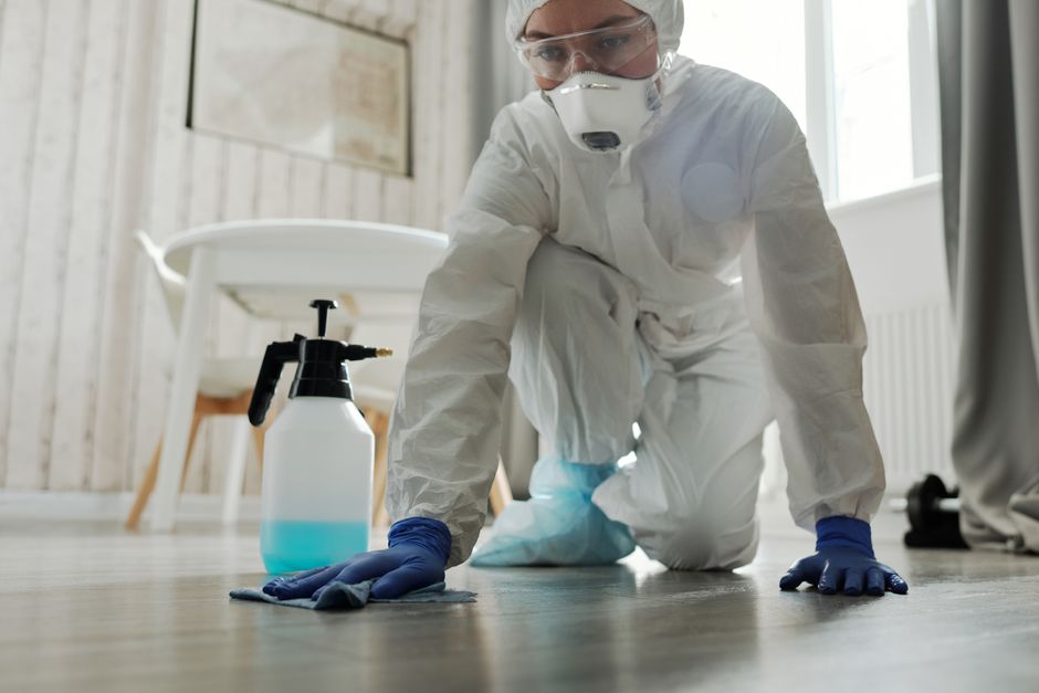 A professional cleaner dressed in white protective suit, safety goggles, and a face mask is kneeling on a wooden floor, using a cleaning cloth to wipe the surface. Nearby, there is a spray bottle fill