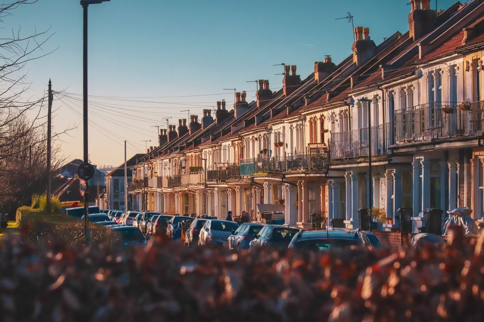 A quiet residential street in Chadwell Heath with brick terraced houses featuring bay windows and ornate chimneys. The street is lined with lush green trees and well-maintained gardens with trimmed he